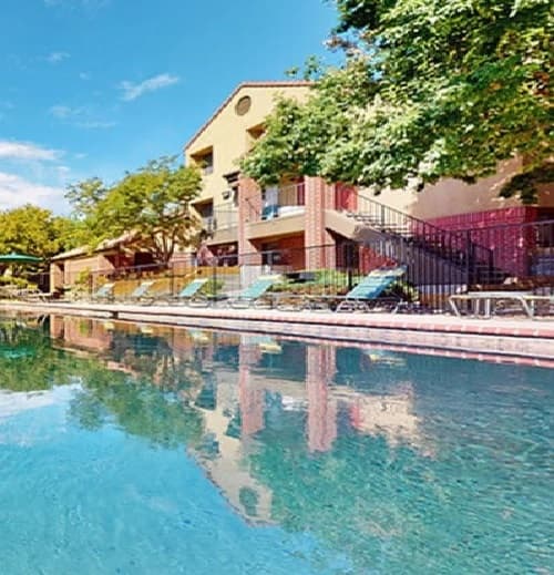 A clear outdoor swimming pool at Ram's Village Fort Collins reflects a multi-story apartment building with balconies, surrounded by trees and several lounge chairs on the pool deck under a bright blue sky.