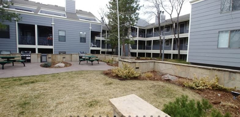 The courtyard at Blue Sky Lofts Boulder features gray apartment buildings with balconies and windows, picnic tables, benches, small trees, and landscaped garden beds filled with grass and shrubs.