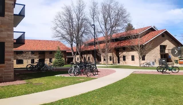 A campus scene with tan-brick buildings, red-tiled roofs, and leafless trees near Smith Hall Boulder. Several bicycles are parked along paths on green grass under a partly cloudy sky.