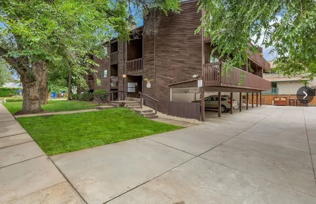 A modern, two-story apartment building with dark wood siding, balconies, a covered parking area with cars, concrete driveways, green lawn, and large leafy trees in front at South Street Apartments Boulder.