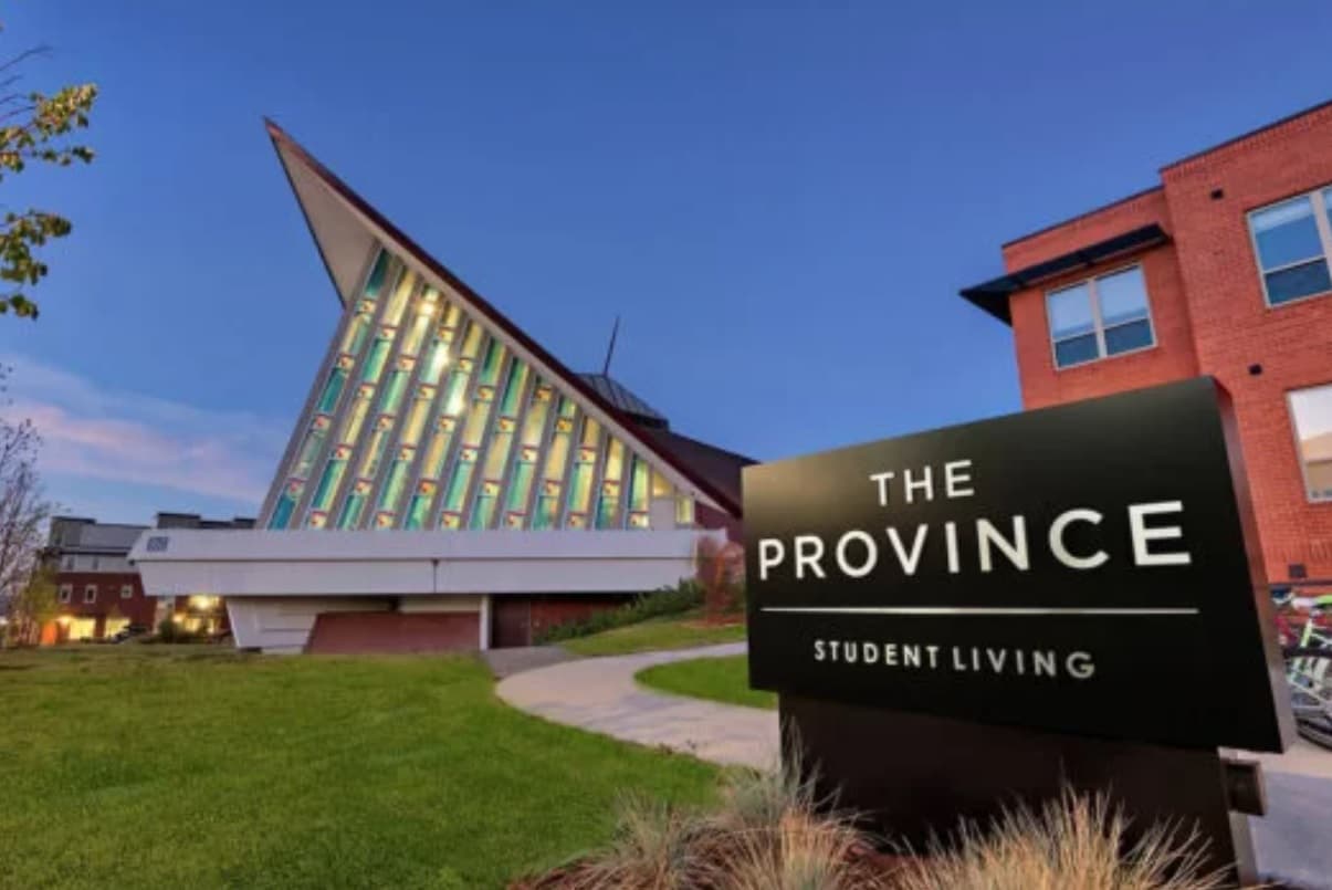 Modern, angular building with colorful vertical windows at sunset, next to a brick building. In the foreground, a black sign reads The Province Boulder Boulder Student Living on a grassy lawn.