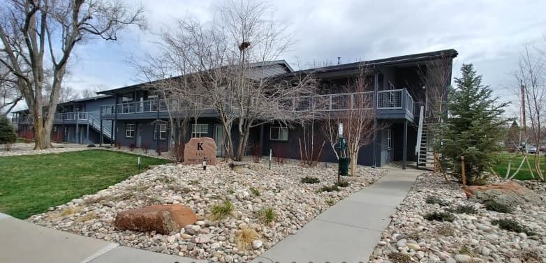 A two-story apartment building with a dark exterior at Timber Ridge Apartment Homes Boulder, surrounded by leafless trees and a rock garden. A sidewalk leads to the entrance, with a stone sign marked "K" near the walkway.