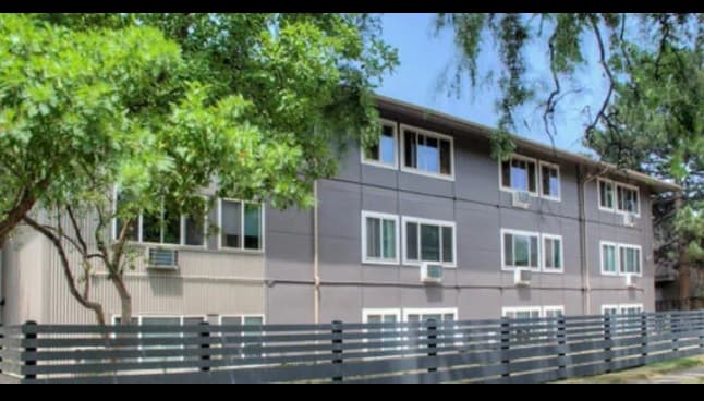 A three-story apartment building with gray siding and white-framed windows, Twin Pines Boulder is surrounded by green trees and a modern horizontal slat fence under a clear blue sky.