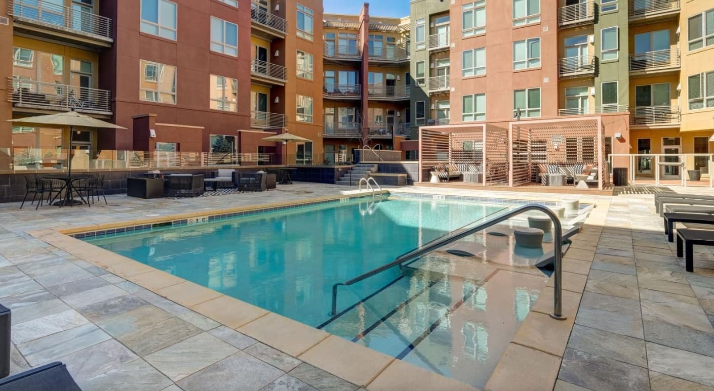 Modern apartment complex courtyard at Two Nine North Boulder featuring a swimming pool, lounge chairs, patio tables with umbrellas, and secure fencing. Multi-story buildings with balconies surround the inviting pool area.