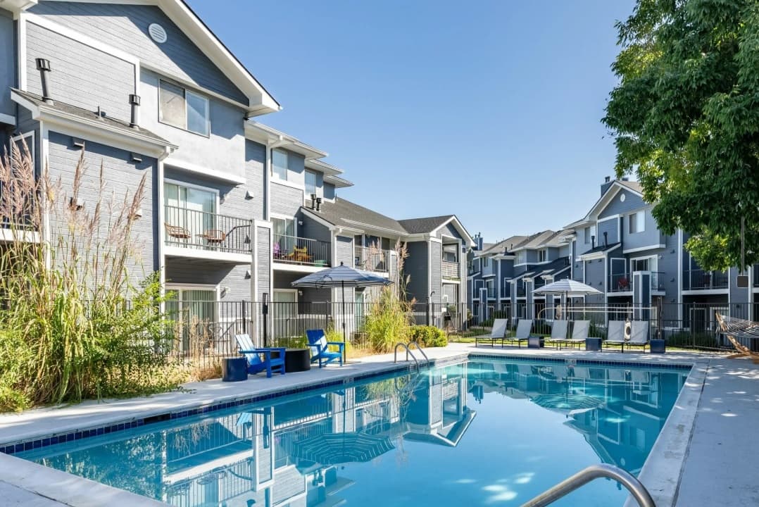 Outdoor swimming pool with clear blue water at Union Baseline Boulder, surrounded by lounge chairs, a gazebo, and modern gray apartment buildings under a clear sky. Green plants and trees enhance the relaxing pool area.