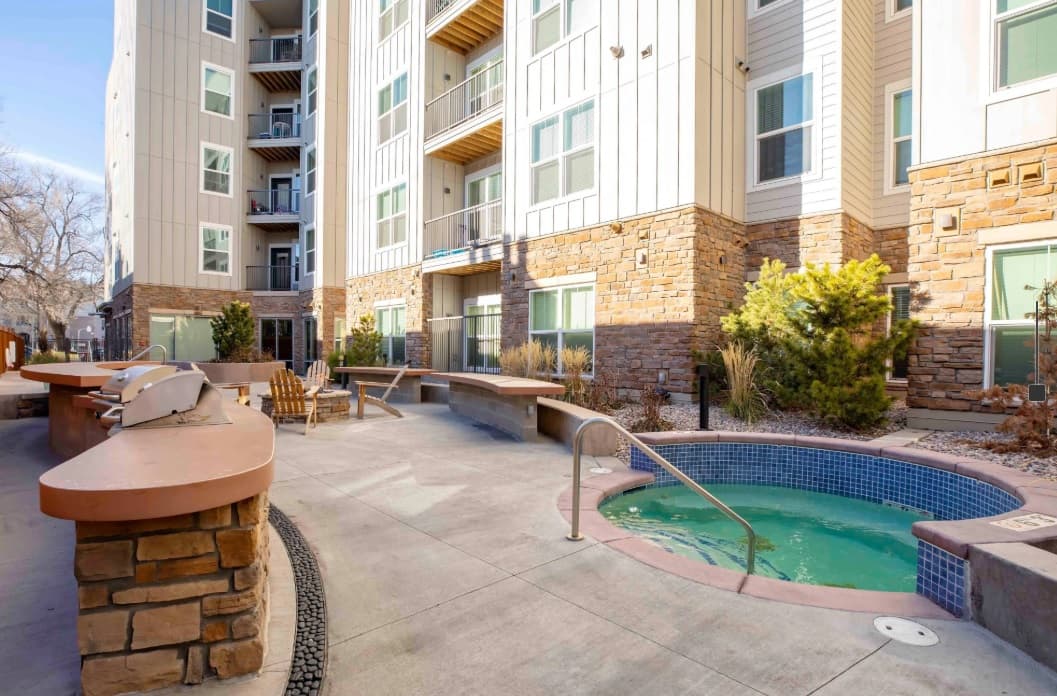 Relax at Union on Plum Fort Collins in the inviting outdoor area, featuring a small round hot tub, built-in barbecue grill, comfortable seating areas, and lush landscaping with shrubs and trees—all surrounded by multi-story buildings.