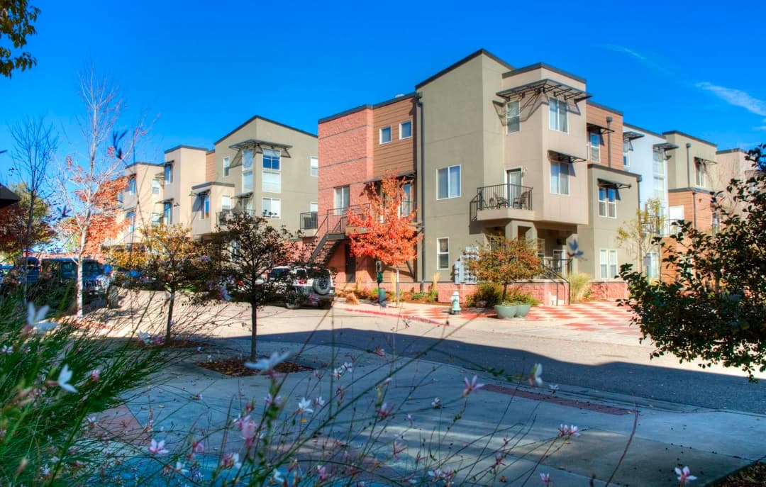 Modern three-story Uptown Broadway Apartments Boulder with balconies and large windows, surrounded by autumn trees and parked cars, under a clear blue sky on a sunny day. Grass and flowers are visible in the foreground.