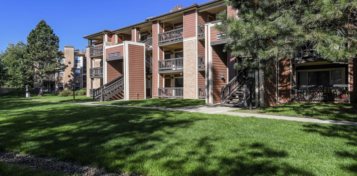 Verdant Apartment Homes Boulder features three-story buildings with wooden balconies and exterior staircases, all surrounded by lush green grass, trees, and a clear blue sky on a sunny day.