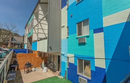 A colorful apartment building, View House Boulder, features a blue geometric mural on its brick wall, a wooden fence, and a small patio with a grill. Trees and another house are visible in the background.