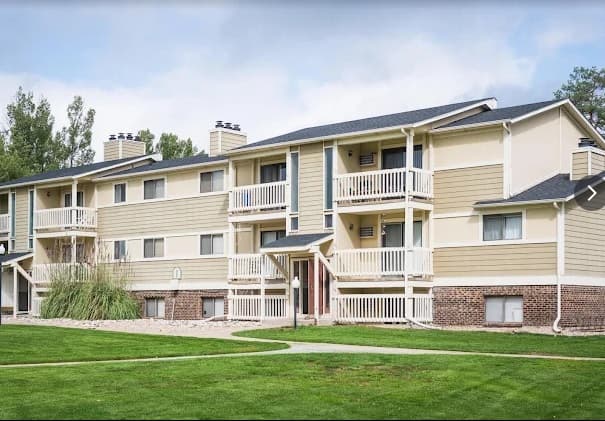 Village Gardens Apartments Fort Collins features a three-story building with beige siding, white trim, balconies, and a brick base, surrounded by green grass and trees under a partly cloudy sky.
