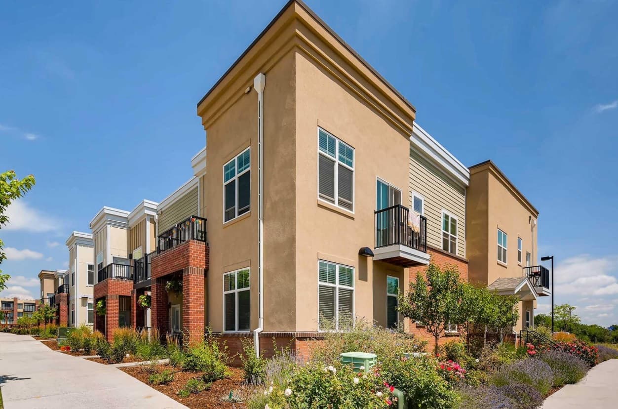 Modern apartment building with tan and brick exterior, multiple balconies, and large windows—Violet on Broadway Apartments Boulder boasts landscaped gardens and a sidewalk under a clear blue sky.