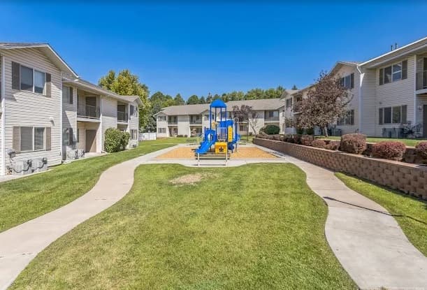 A small playground with a blue and yellow play structure sits in the center of Washington Park Apartments Ogden, surrounded by lawns, sidewalks, and beige two-story apartment buildings under a clear blue sky.