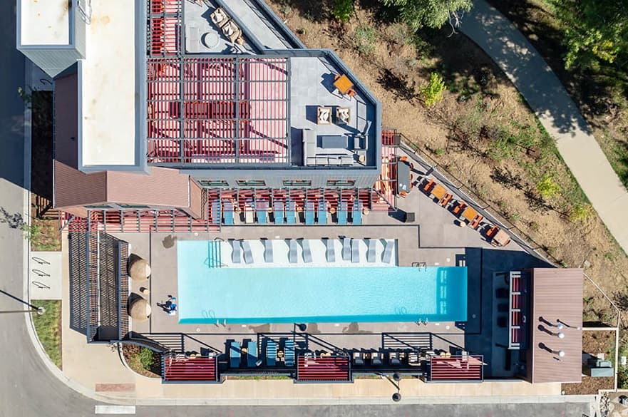 Aerial view of the modern outdoor swimming pool at Weathervane Apartments Boulder, with lounge chairs on both sides, shaded seating areas, and landscaped surroundings. The rectangular pool is bordered by buildings and pathways.