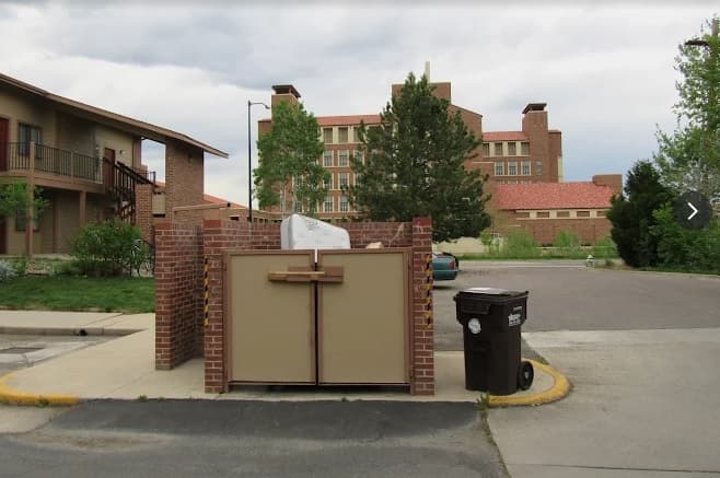 A gated trash enclosure with tan doors and brick walls stands beside a recycling and trash bin near the street at Wimbledon Apartments Boulder, with apartment buildings and trees visible in the background.