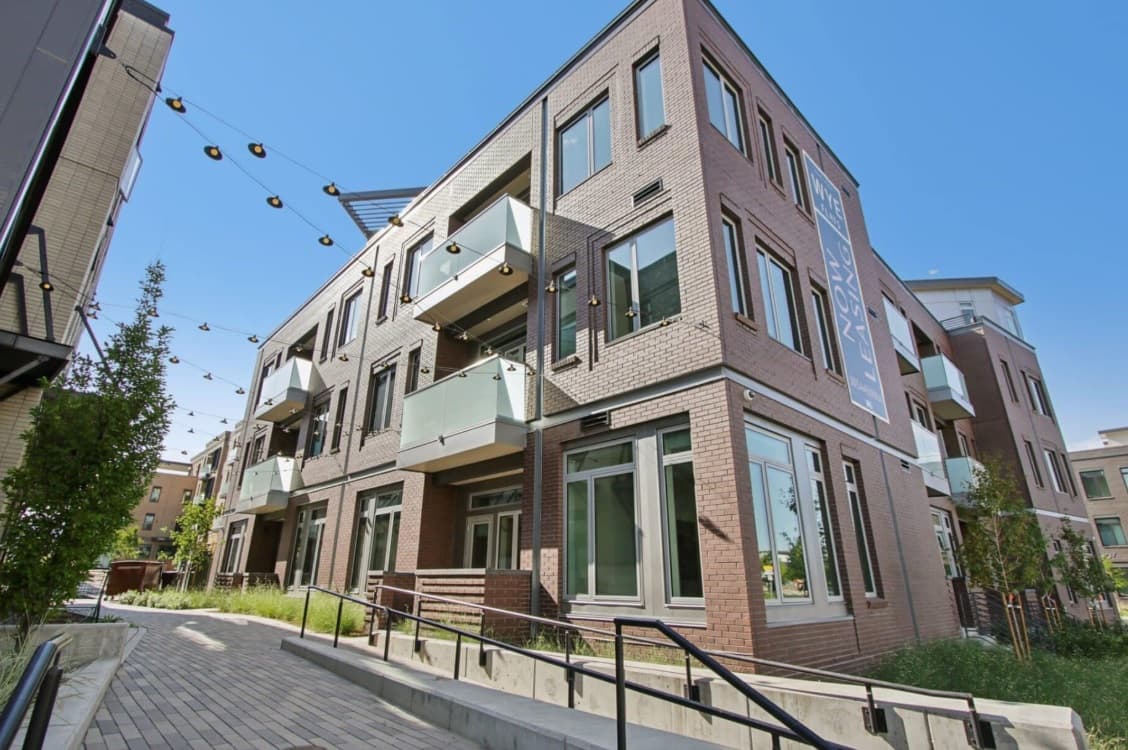 Modern three-story apartment building, Wye Flats Boulder, features a brick exterior, large glass windows, and small balconies. String lights hang above a paved walkway lined with greenery and railing under a clear blue sky.