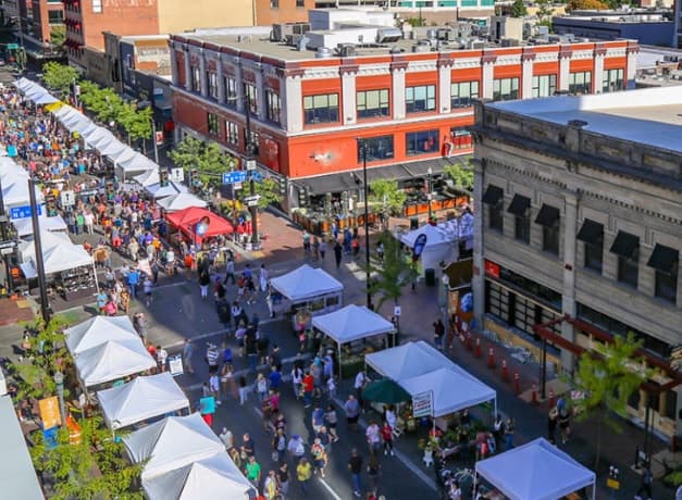 Aerial view of a busy street festival with white tents, booths, and crowds of people between two multi-story buildings in Boise’s Ascent East End on a sunny day.
