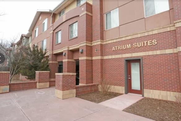 A three-story brick building with a sign reading Atrium Suites Fort Collins above the door, featuring large windows and a paved walkway in front. Some small trees and shrubs are near the entrance.