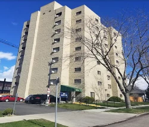 The Bannock Arms Apartments Boise is a multi-story beige apartment building with several windows, a green awning over the entrance, cars parked in front, a leafless tree nearby, and a clear blue sky in the background.