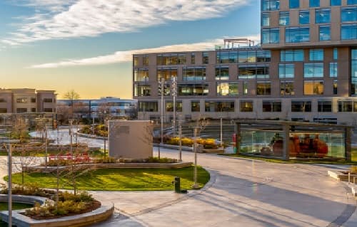 Modern office buildings with glass windows surround a landscaped courtyard near Ascent BoDo Apartments Boise, featuring curved walkways, green lawns, benches, and small trees under a partly cloudy sky at sunset.