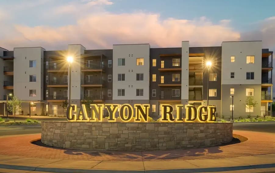Modern apartment complex at sunset with illuminated Canyon Ridge Apartments Boise sign in front, surrounded by a low stone wall and neatly landscaped area.