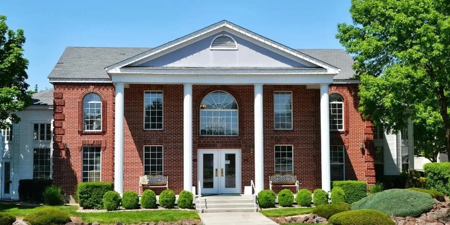Carriage Crossing Apartments Boise features a large red-brick building with tall white columns, large arched windows, and neatly trimmed bushes and trees in front, all beneath a clear blue sky.
