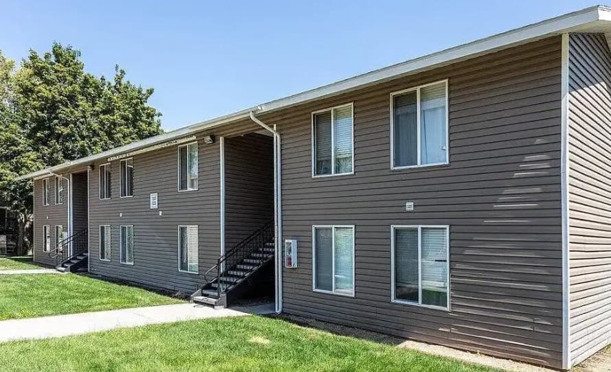 Two-story Central Pointe Boise apartment building with brown siding, multiple windows, and two black metal staircases leading to the upper level. The building is surrounded by green grass and trees under a clear sky.