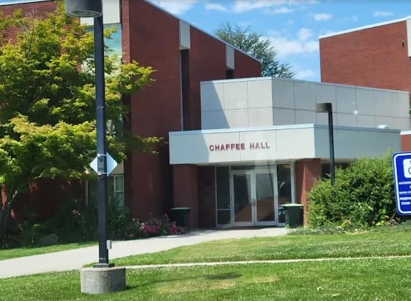 A brick building labeled Chaffee Hall Boise at its entrance, surrounded by green grass, bushes, a tree, a lamp post, and a blue accessible entrance sign on the right. The sky is partly cloudy.