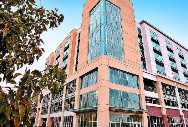 A modern mid-rise building with large glass windows and tan-colored exterior walls at Civic Plaza Boise, viewed from a low angle, with a leafy tree in the foreground and a clear blue sky above.