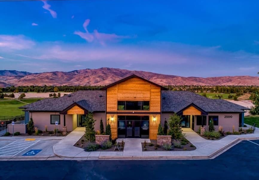 A modern, single-story building with wood accents is pictured at sunset in Columbia Village Boise, set against rolling hills and blue sky. The entrance is well-lit, and there is a visible accessible parking spot in front.