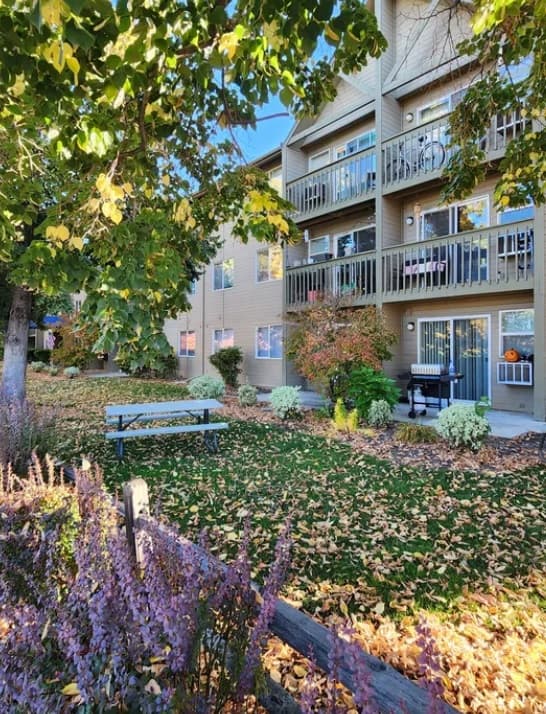 The Conservatory Boise features a three-story apartment building with balconies, surrounded by autumn trees and fallen leaves. A picnic bench sits on the lawn, edged by flower beds with purple and white plants across the green grass.