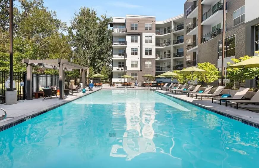 Outdoor swimming pool surrounded by lounge chairs, umbrellas, and seating areas at Cortland on the River Boise, with a modern multi-story apartment building and trees in the background under a clear sky.
