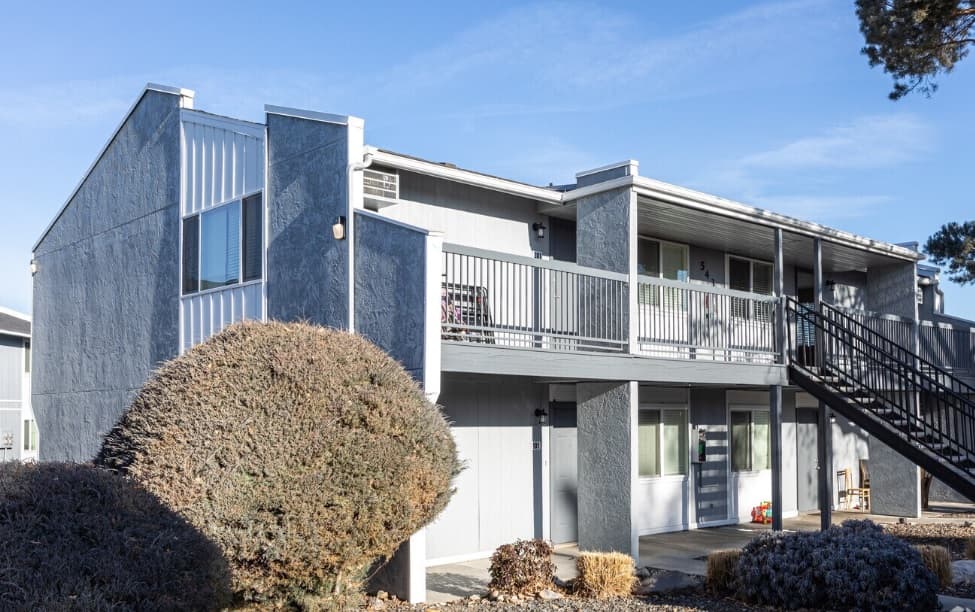 Curtis Junction Apartments Boise is a two-story apartment building with a gray exterior, outdoor stairway, and metal railings. Well-maintained bushes and stone landscaping are visible in front under a clear blue sky.
