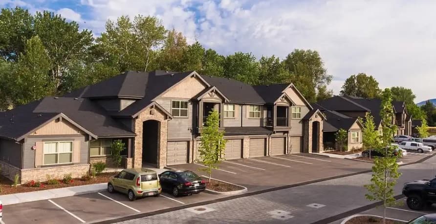 The District at Parkcenter Boise is a modern apartment complex with tan siding and brick accents, featuring attached garages and a parking lot with several parked cars, surrounded by trees under a partly cloudy sky.
