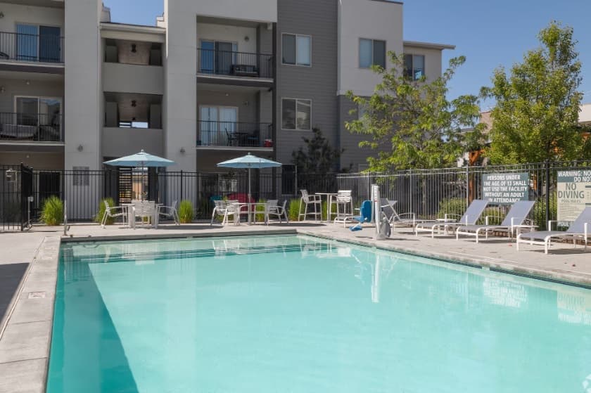 The outdoor swimming pool at Encore Skyline Luxury Apartments Boise is surrounded by lounge chairs and tables with blue umbrellas, all within a secure fence. Apartment balconies overlook this inviting space for relaxation and leisure.