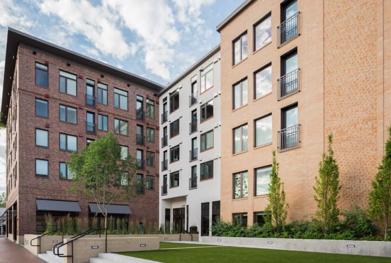 A modern apartment complex at 5th & Idaho Boise, featuring brick and white facades, large windows, small balconies, landscaped greenery, and a lawn area in the foreground under a partly cloudy sky.