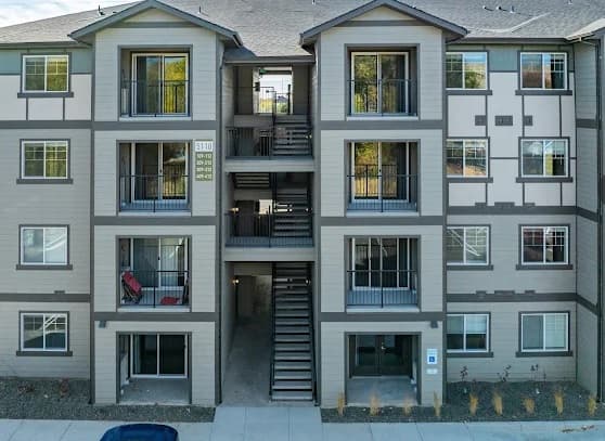 Haven on State Boise is a three-story apartment building with a gray and white exterior, balconies, large windows, and a central staircase. A few chairs rest on the balconies, complemented by attractive landscaping in front.