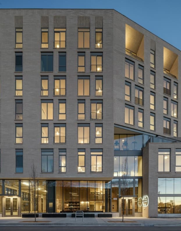 Modern multi-story building with large windows illuminated from inside at dusk. The facade features a mix of glass and light-colored brick. Hearth on Broad Boise is visible amid bare trees and a bike rack along the sidewalk out front.