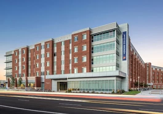 A modern, multi-story brick and glass building labeled Honors College stands next to Sawtooth Hall Boise, alongside a road with blurred light trails from passing vehicles, set against a clear sky at dusk.