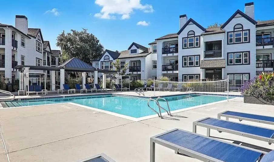 Enjoy the outdoor swimming pool at Huntington Apartments Boise, surrounded by blue lounge chairs and a shaded seating area, with modern three-story apartment buildings in the background under a bright blue sky.