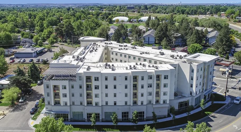 Aerial view of Identity Boise, a large, modern white apartment building with several floors, surrounded by trees and residential houses under a clear blue sky. Streets and parked cars are visible around the building.