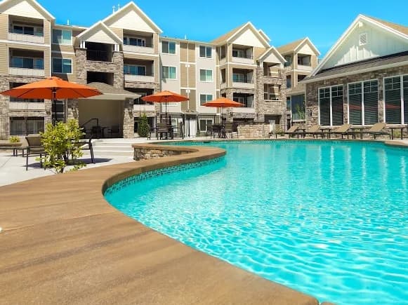 Modern apartment complex, Kensington at North Pointe Boise, features a large outdoor swimming pool, lounge chairs, and orange umbrellas, set against multi-story buildings with stone and beige siding under a clear blue sky.