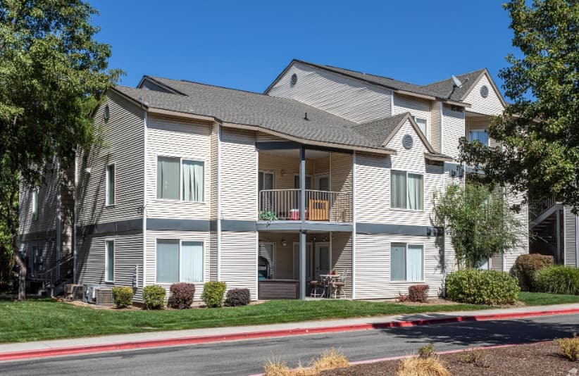 A two-story beige apartment building with balconies and large windows, Liberty Lake Apartments Boise is surrounded by green grass, shrubs, and trees under a clear blue sky.