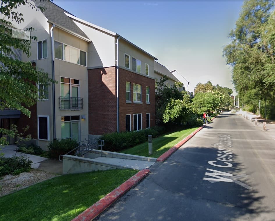 A street view of a modern red-brick and tan apartment building near Morrison Hall Boise, with balconies, trees, and greenery along the sidewalk. The street is labeled “W Cesar E Chavez Blvd.” The sky is clear and sunny.
