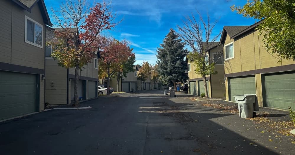 A quiet residential street lined with modern Monterra Townhomes Boise, each featuring a double garage. Trees with autumn leaves border the roadway, and a few trash bins are visible on the asphalt under a clear blue sky.