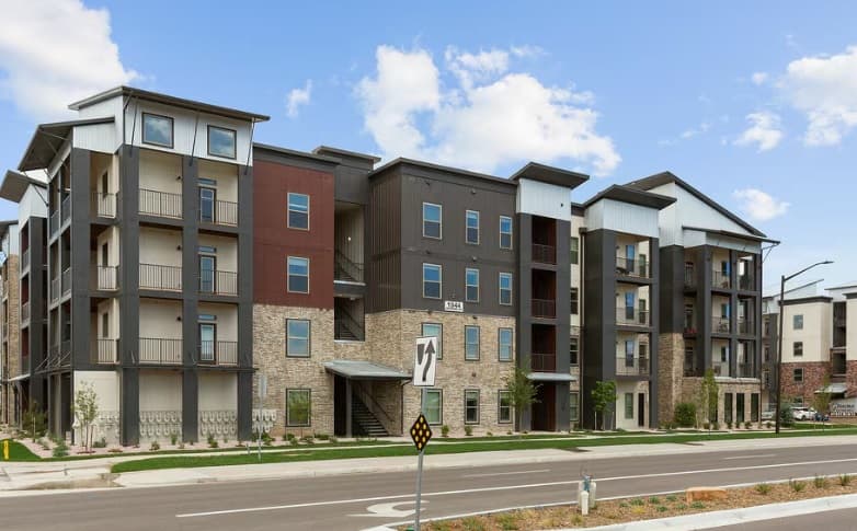 Modern apartment complex, Crowne at Old Town North Fort Collins, with multiple stories, balconies, and a mix of brick and colored siding. The building faces a paved road with a sidewalk, landscaped areas, and a clear blue sky above.