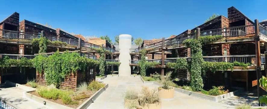 A courtyard at Plaza Apartments Boise features lush green plants and vines draping over wooden structures, surrounded by two-story buildings with balconies and a cylindrical tower in the center beneath a clear blue sky.