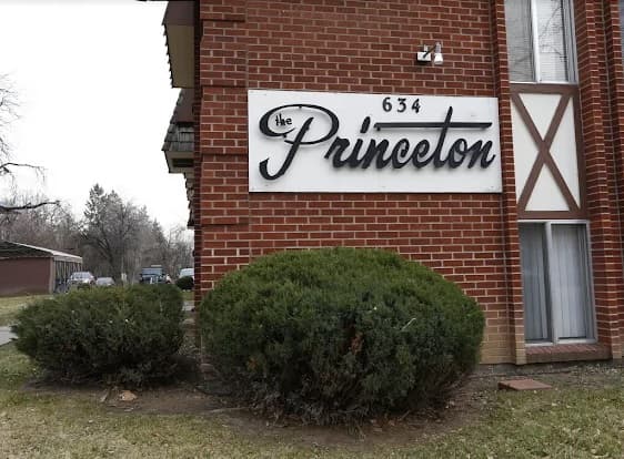 A brick apartment building with a white sign that reads the Princeton 634 in black script font. Two bushes are in front of The Princeton Fort Collins, with a window visible on the lower right side.