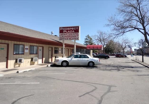 A light-colored car is parked in front of Prospect Plaza Apartments Fort Collins, a one-story building with a sign for the Laundromat and Office Suites. Several doors and windows are visible, with trees lining the mostly empty parking lot.
