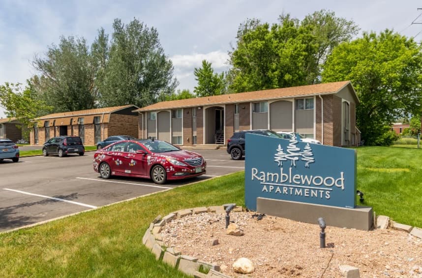 A blue sign reading Ramblewood Apartments Fort Collins stands in front of a parking lot with cars and a brick apartment building surrounded by green trees and grass on a sunny day.