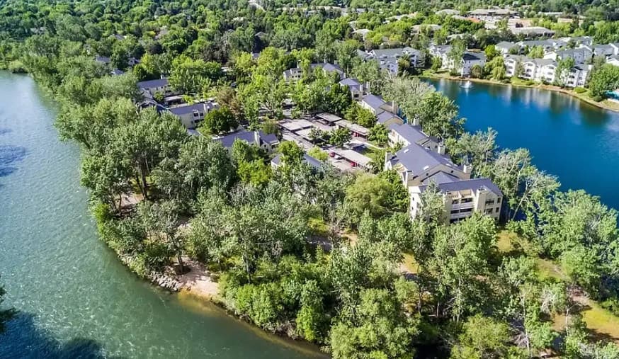 Aerial view of Reedhouse Apartments Boise, an apartment complex surrounded by dense trees, nestled between a river and a lake, with lush greenery and nearby residential buildings in sight.